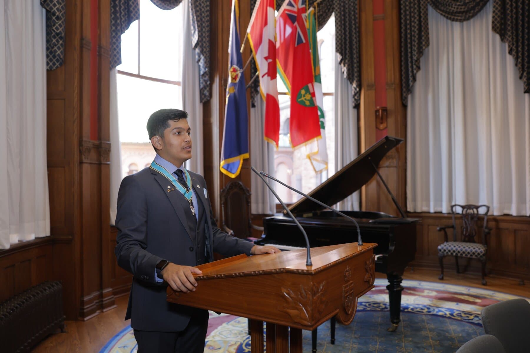 Adil Mukhi speaking at the podium at Queen's Park, wearing the Ontario Medal for Young Volunteers